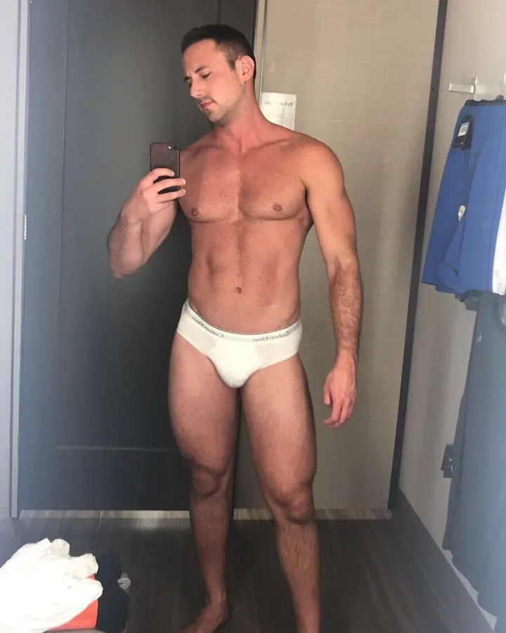 Homme torse nu en sous-vêtements blancs, prenant un selfie dans une pièce moderne.