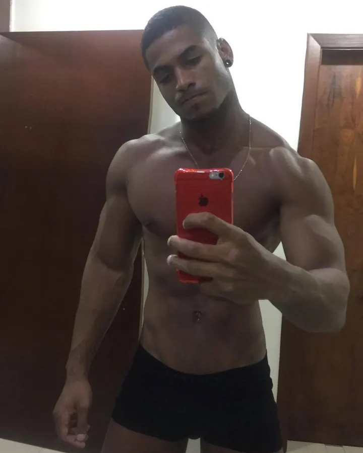 Homme torse nu, peau mate, regard intense, tenant un téléphone rouge devant lui, souriant légèrement, en sous-vêtements noirs.