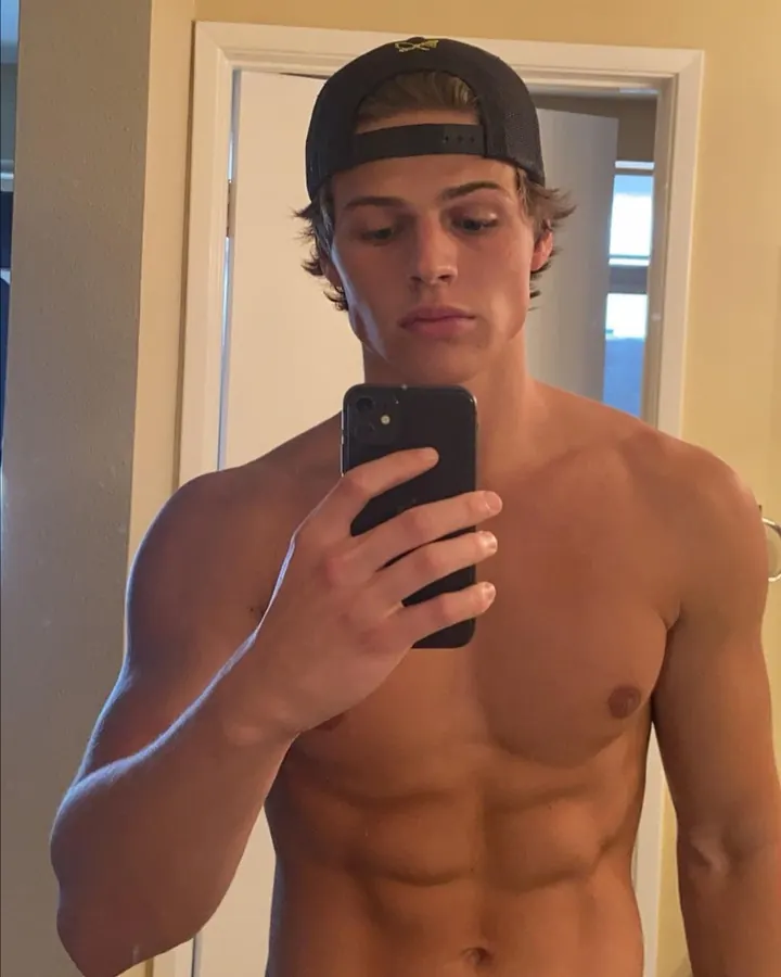Jeune homme torse nu, portant un t-shirt blanc à manches courtes, tirant le tissu pour exposer son torse, regard vers le côté, pantalon noir avec bande Calvin Klein.