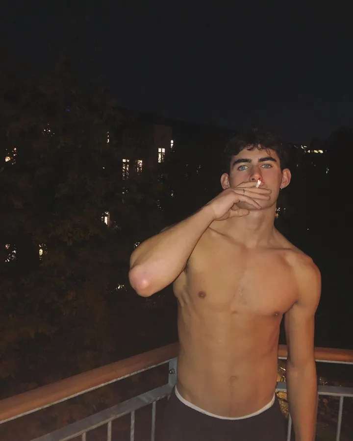 Jeune homme torse nu, portant un short noir, allongé sur une serviette violette, entouré de tatouages complexes, regardant vers le haut, en plein air avec vue sur bâtiments.