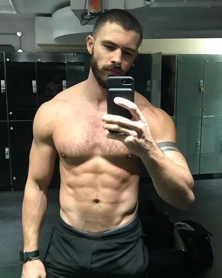 Homme européen en t-shirt gris, barbe courte, prenant un selfie dans un miroir, regard concentré, écouteurs blancs, lumière neutre.