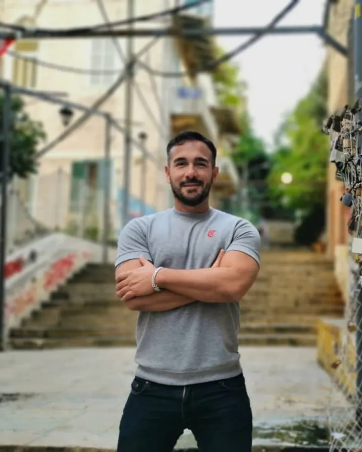 Homme européen, barbe, t-shirt gris, jeans, debout en ville, bras croisés, sourire discret, fond flou avec escaliers et cadenas.