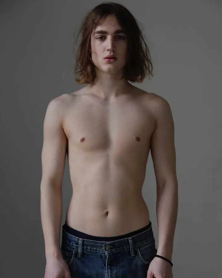 Jeune homme torse nu, peau blanche, cheveux châtains mi-longs, regard direct, jeans bleus, bracelet noir.