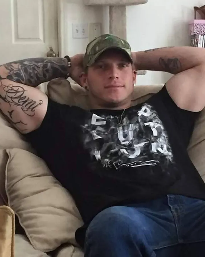 Homme torse nu, peau blanche, tatouages aux bras, casquette camouflage, t-shirt noir 'Fury', allongé sur un canapé.