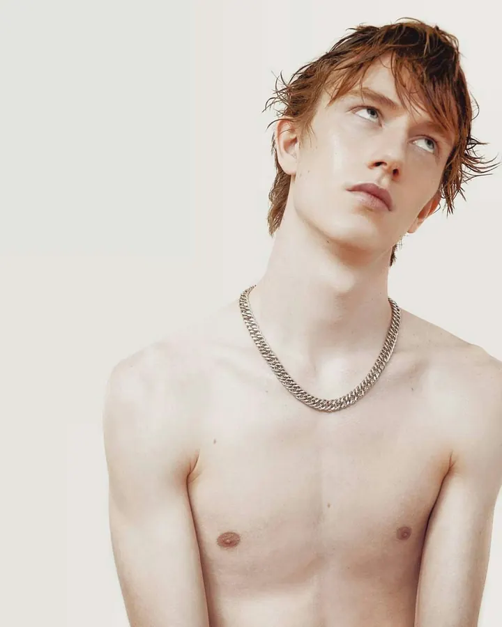 Jeune homme torse nu, peau blanche, cheveux blonds humides, collier en chaîne, regard vers le haut.