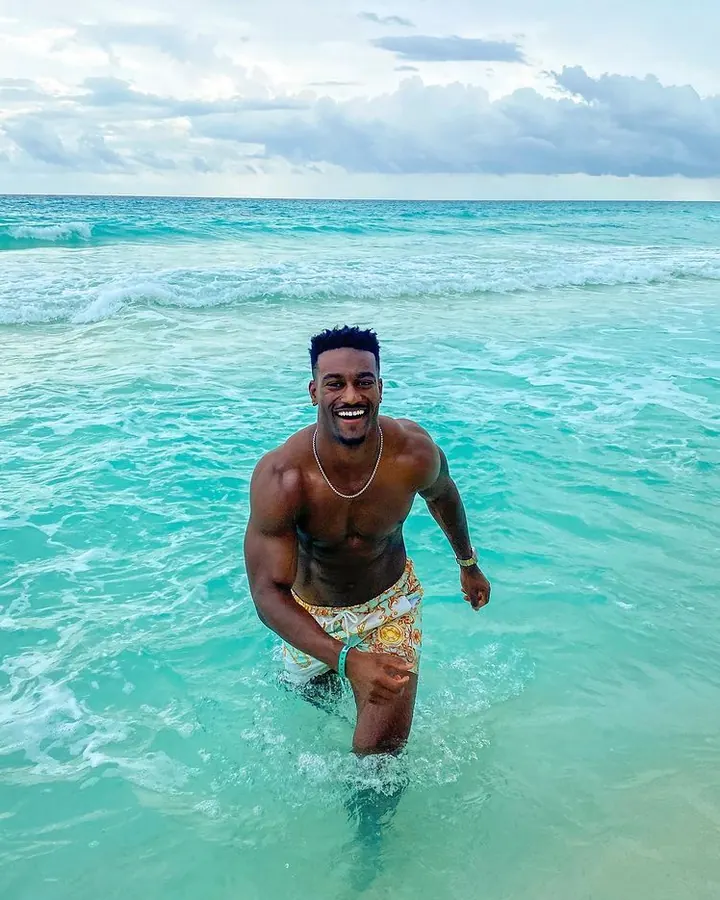 Homme torse nu, peau noire, souriant largement, dans l’eau turquoise, short coloré, collier argenté.