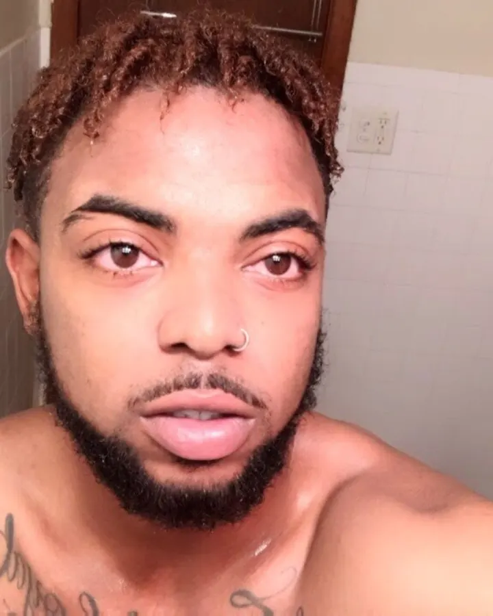 Homme torse nu, peau foncée, barbe courte, regard direct, tatouages sur la poitrine, devant un carrelage blanc.