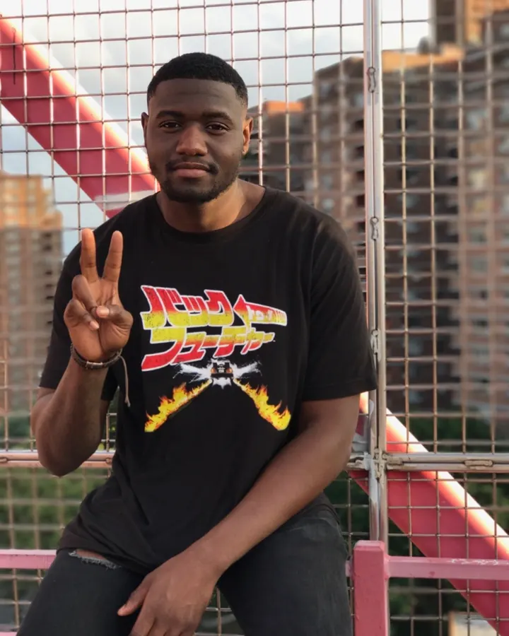 Homme noir en t-shirt noir avec graphisme japonais, assis sur une structure métallique rose.