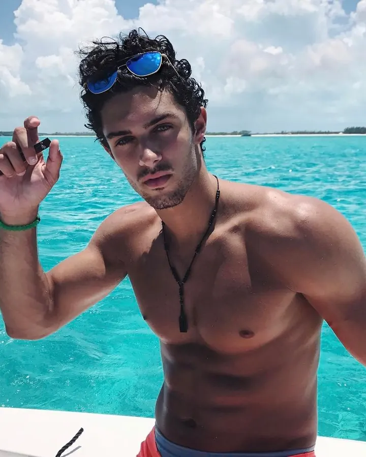 Homme torse nu, barbe courte, regard intense, en plein air, mer turquoise en arrière-plan, lunettes de soleil sur la tête.