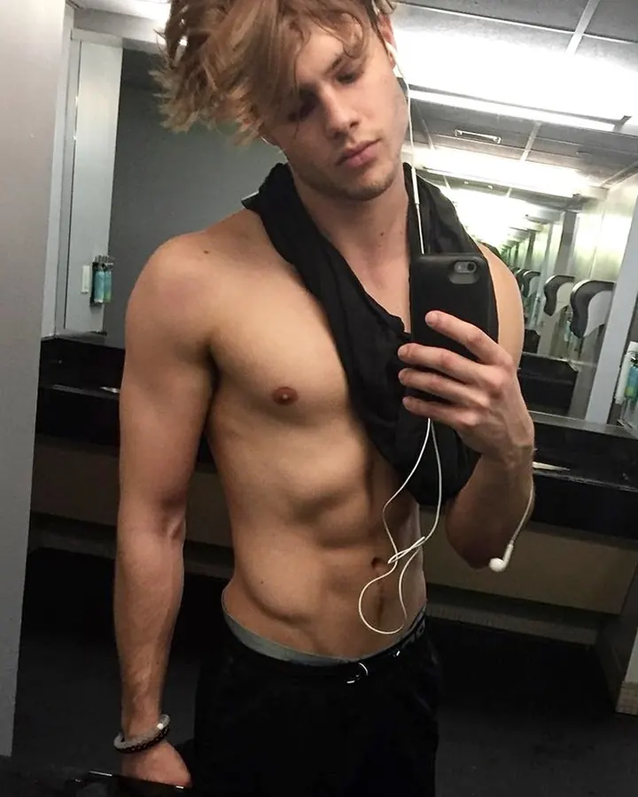 Homme torse nu aux cheveux blonds, prenant un selfie devant un miroir dans une salle de bain, avec un téléphone et des écouteurs blancs.