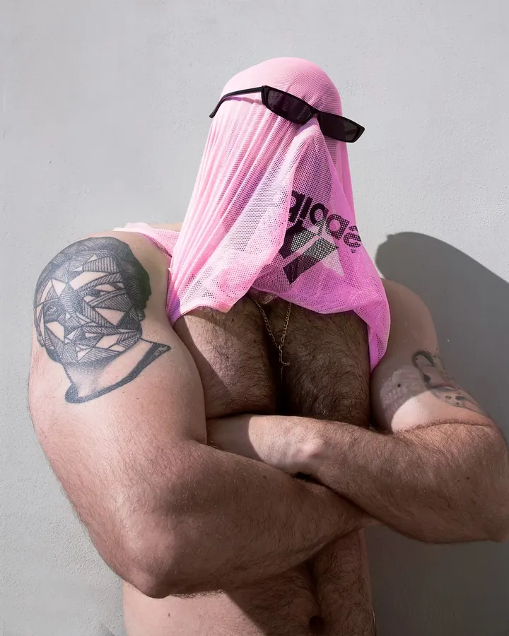Homme musclé avec un tissu rose sur la tête et des lunettes de soleil, bras croisés, tatouage visible sur l’épaule