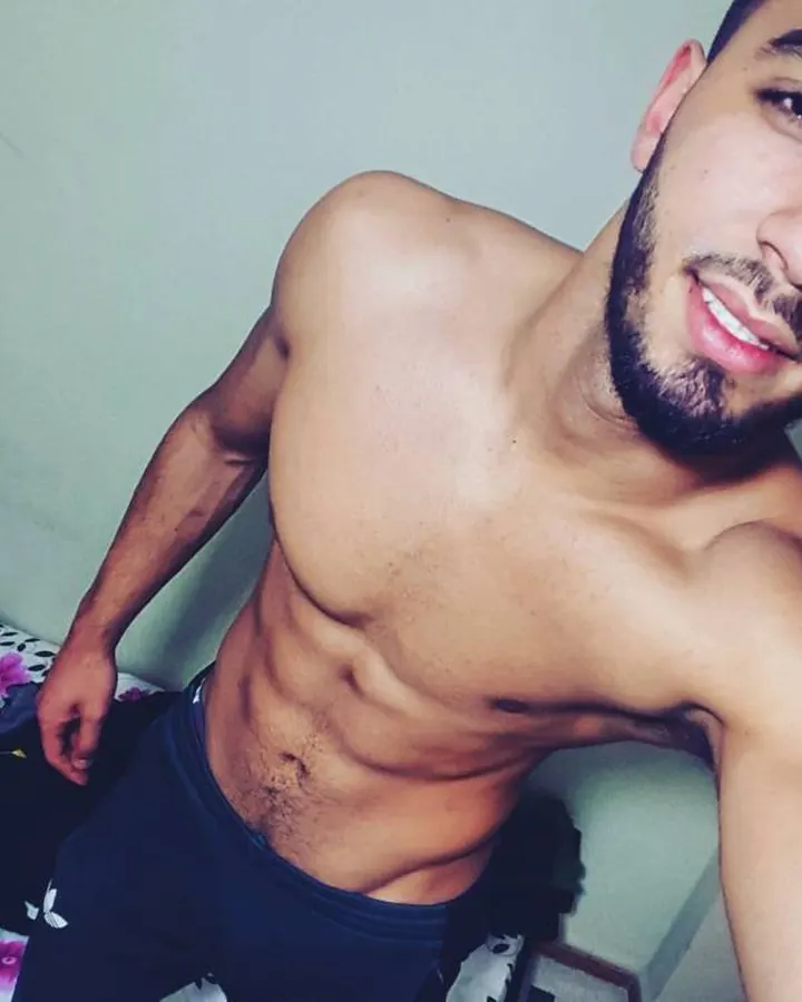 Homme torse nu, souriant, en sous-vêtements, dans une chambre, regard direct vers l'appareil.