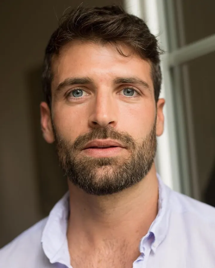 Homme barbu, regardant vers le côté, portant une chemise blanche légèrement ouverte, devant une fenêtre.