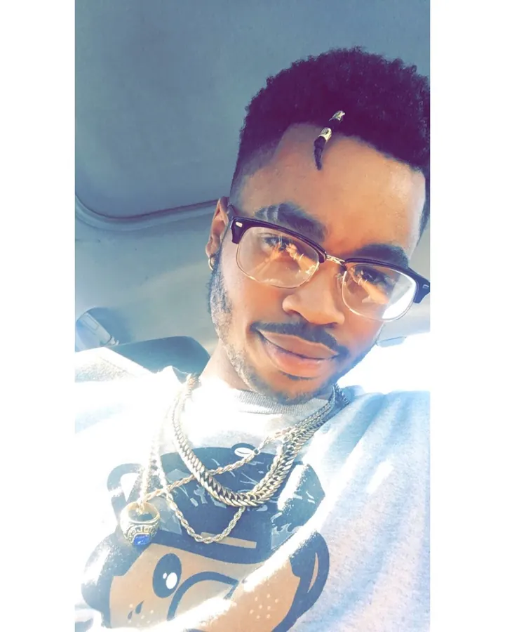 Homme africain aux cheveux courts, portant un t-shirt noir et une chaîne dorée avec une croix, assis dans une voiture.