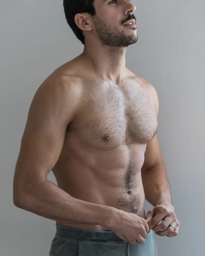 Homme torse nu, aux cheveux bruns noirs, regardant vers le haut, mains posées sur le bas du ventre, devant un fond gris neutre.