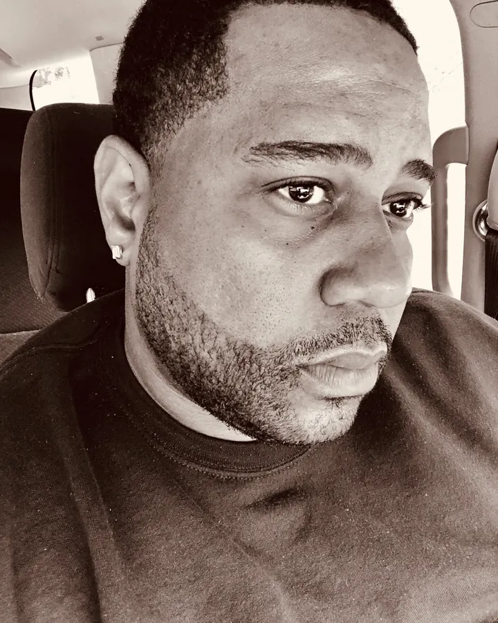 Homme africain aux cheveux courts, barbe courte, regard intense, portant un t-shirt sombre, assis dans une voiture en noir et blanc.