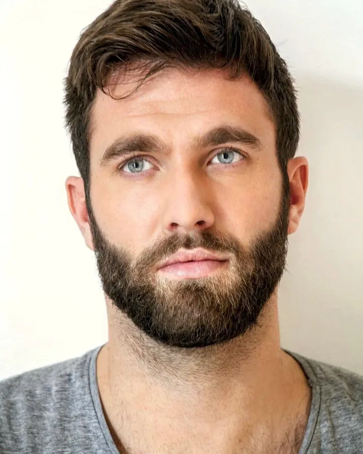 Homme européen aux cheveux bruns-noirs et barbe, torse nu, regardant vers le côté, dans une lumière vive, en plan rapproché.