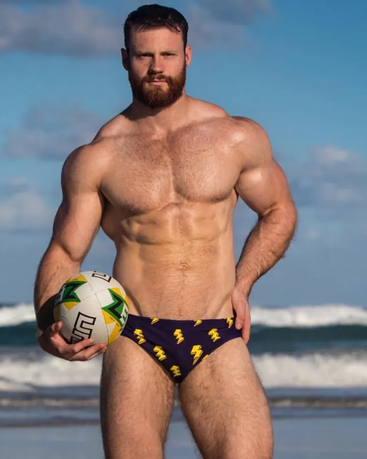 Homme européen aux cheveux bruns-noirs et barbe rousse, torse nu, tenant un ballon de rugby, sur une plage avec des vagues en arrière-plan.