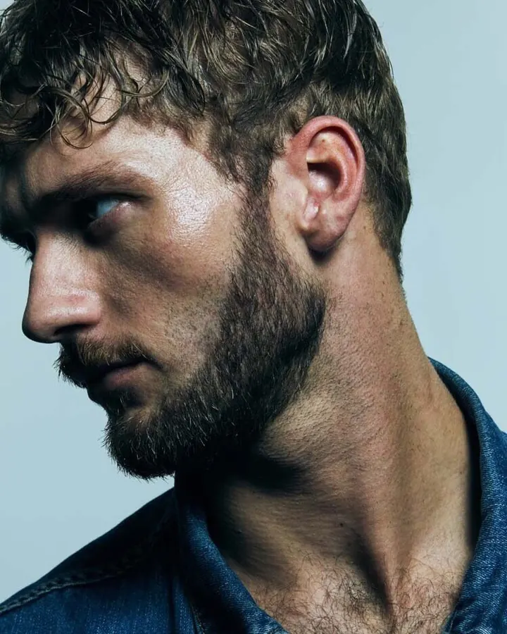 Homme aux cheveux bruns-noirs et barbe, regard intense, vêtu d'une chemise en jean, en plan rapproché sur le profil.