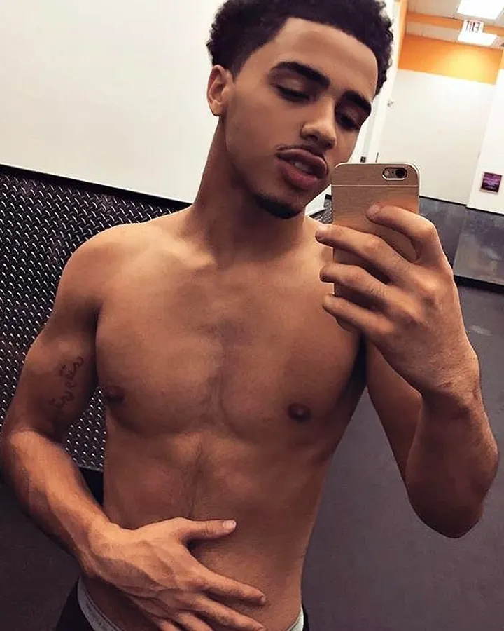 Homme africain de 25 à 35 ans, torse nu, regardant son téléphone, tatouage sur le bras gauche, en salle de sport.
