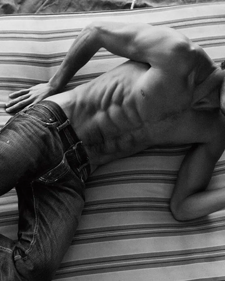 Homme européen de 25 à 35 ans, torse nu, portant un short noir, souriant, dans une pièce sombre.
