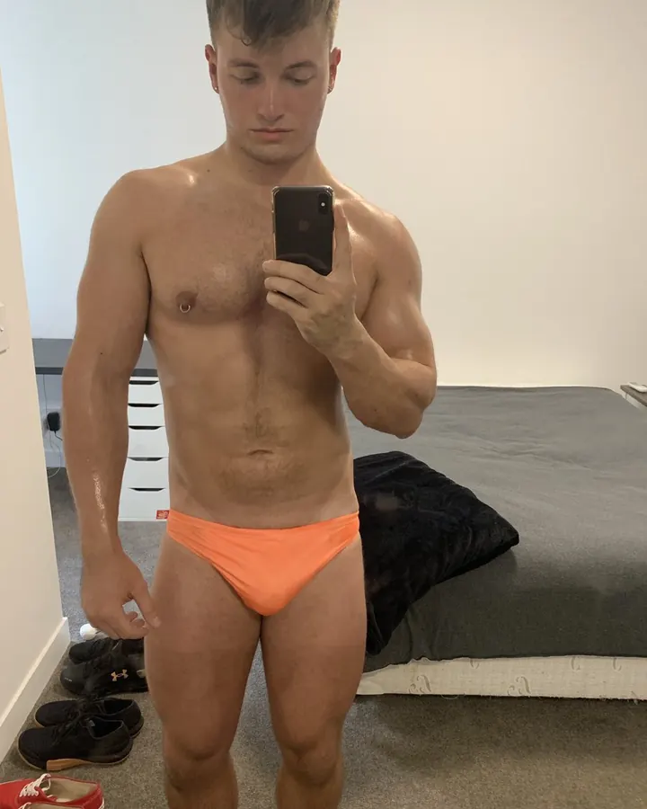 Homme européen torse nu, musclé, en sous-vêtements orange, prenant un selfie devant un miroir dans une chambre.