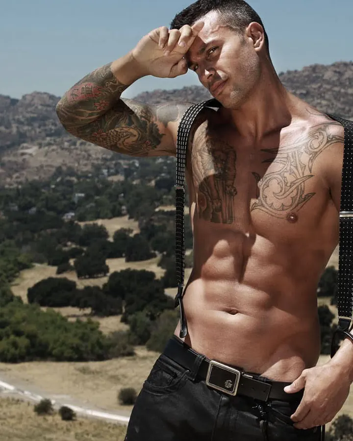 Homme torse nu, debout, regardant vers le haut, avec des suspendeurs noirs, devant un paysage montagneux.