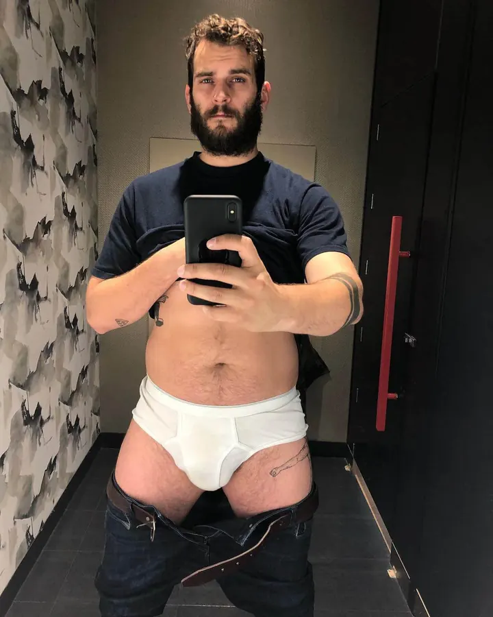 Homme barbu en t-shirt noir, sous-vêtements blancs et jeans, prenant un selfie devant un miroir.