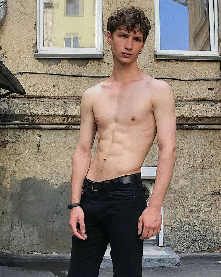 Jeune homme européen aux cheveux blonds, torse nu, debout devant un mur extérieur, regardant l'appareil photo avec assurance.