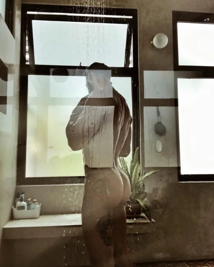 Silhouette d'un homme nu sous la douche, vue de dos, dans une cabine moderne avec des carreaux de verre et une plante verte.