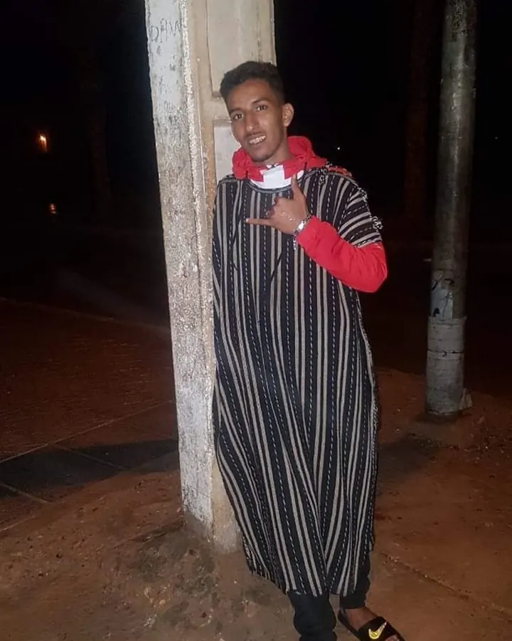 Homme souriant, portant une robe à rayures noires et blanches, un sweat rouge, et des sandales Nike, posant la nuit contre un poteau.