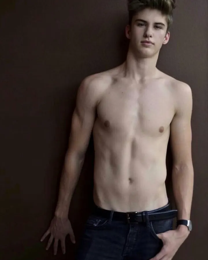 Jeune homme torse nu, aux cheveux clairs, debout contre un mur sombre, portant un jean et une ceinture noire.