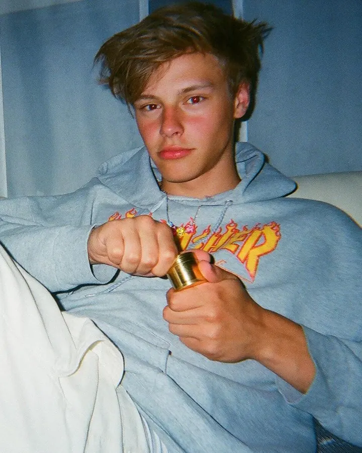 Jeune homme assis sur un canapé, portant un sweat gris à capuche avec un motif flamboyant, tenant un briquet doré.