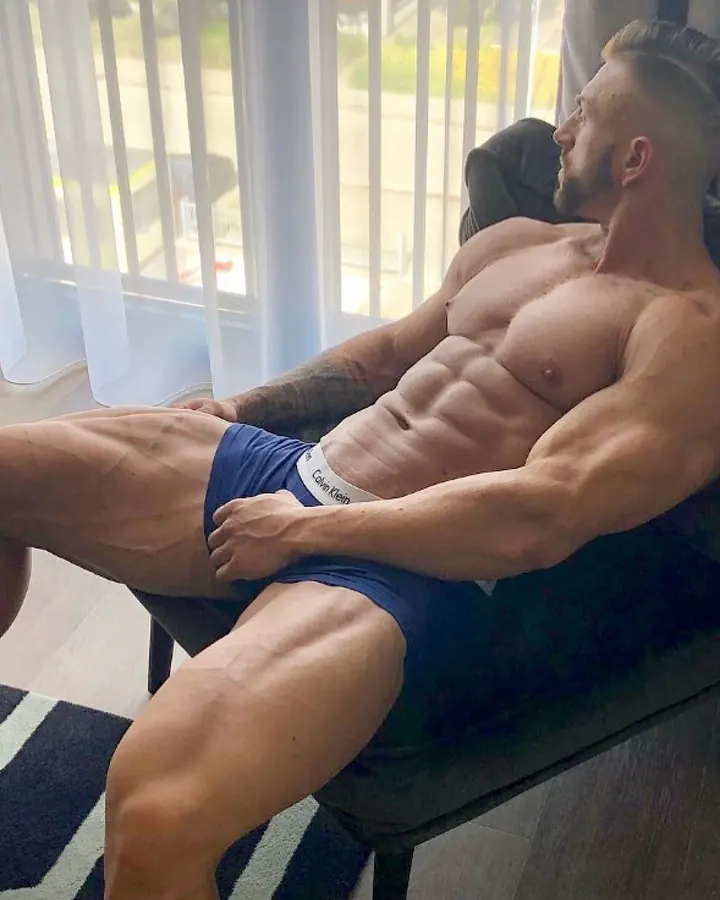 Homme torse nu, musclé, en sous-vêtements bleus, assis sur un fauteuil près d'une fenêtre avec voilages blancs.