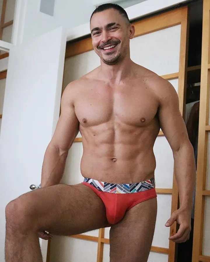 Homme torse nu, souriant, portant un slip rouge à motifs colorés, posant devant une porte japonaise en bois et papier