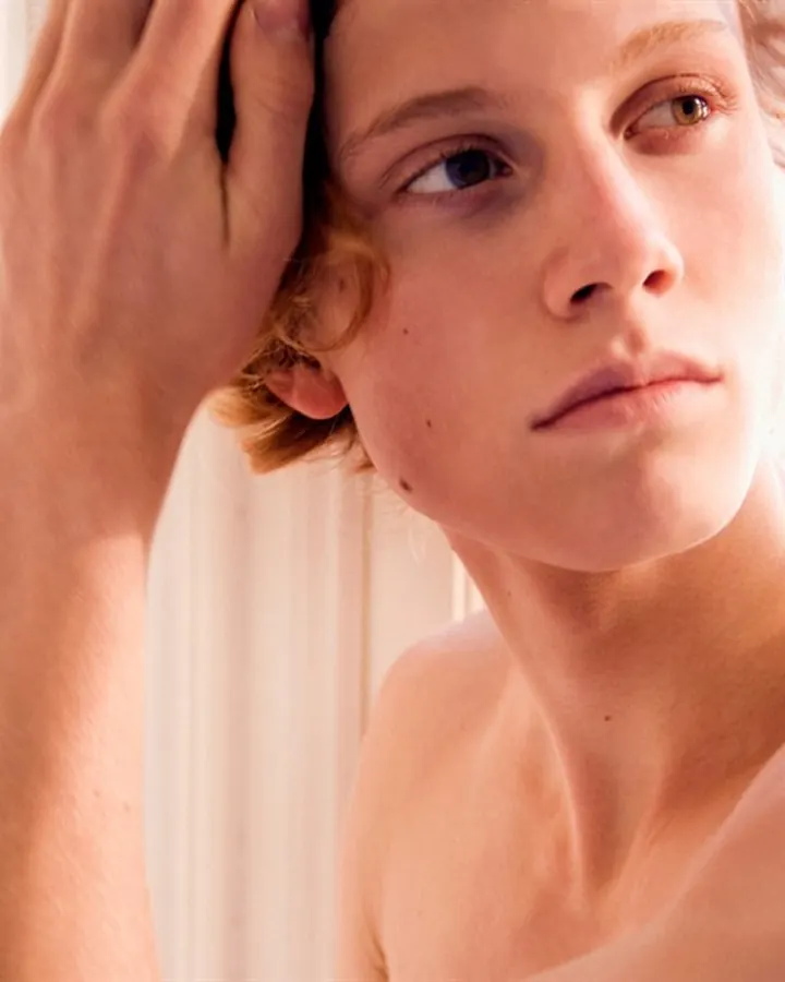 Jeune homme torse nu, aux cheveux blonds, regardant vers le haut, main sur la tête, lumière vive derrière lui